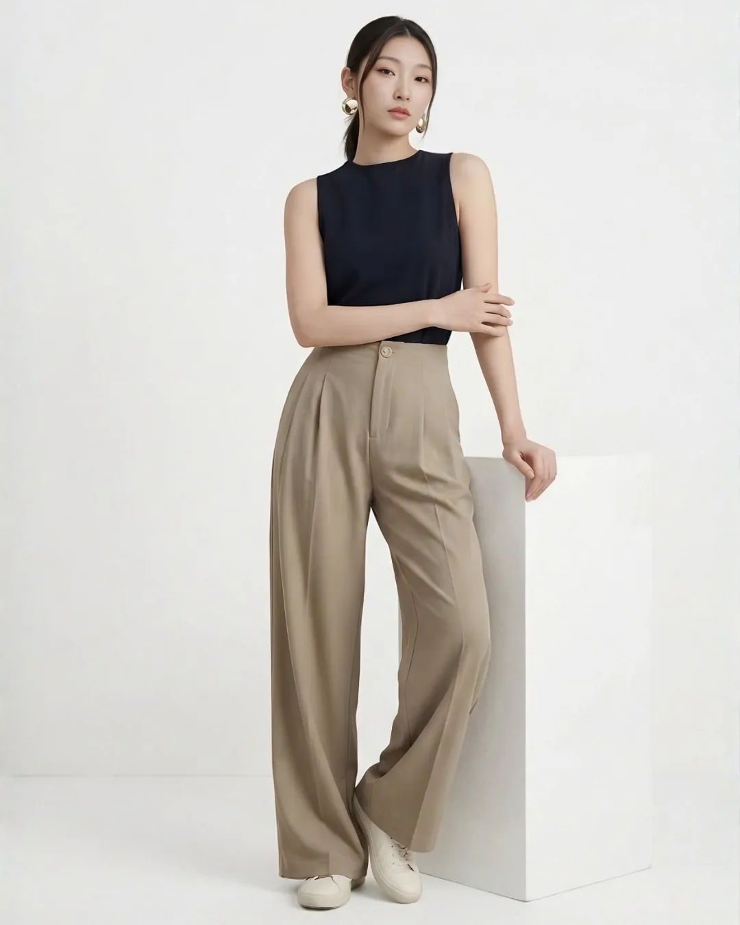 Model memakai celana wide leg krem lipit dan atasan hitam tanpa lengan, bersandar di balok putih